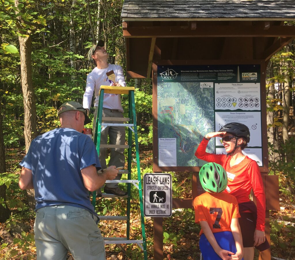 two men work on kiosk, one on a ladder. A woman and boy with bike helmets on stand asking man in blue shirt some questions.