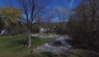 a view of a park with a flowing dirt path called a pump track, rolling up and down making waves over the grass. A mini excavator works, and there is someone on the track.