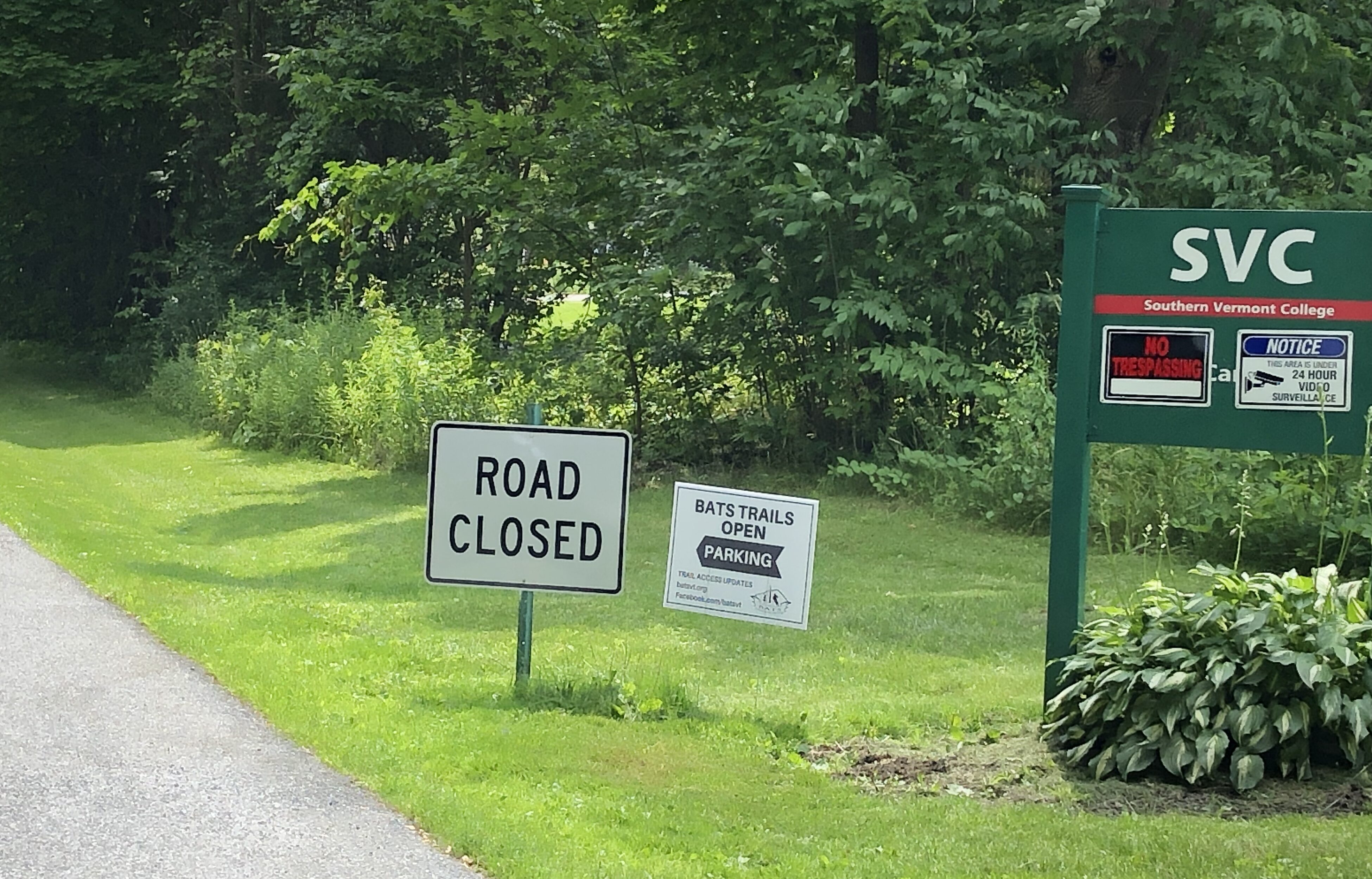 road on the left with signage for Southern Vermont College. One sign reads, "road closed". Second reads "BATS TRAIL OPEN" with left arrow reading "parking" and the url www.batsvt.org, the third on the right is SVC sign with a "No Tresspassing Sign" and a security/cameras on the property sign.
